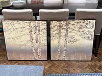 Pair of framed prints showing birch trees side by side on the floor in front of dining chairs and table, highlighting full view of artwork and frames.