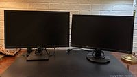 Both monitors side by side on a black surface with visible power cables and stands, against a white brick wall.
