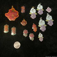 Top-down view of all enamel charms arranged on black fabric with a US quarter coin for scale. Shows variety of colorful floral enamel pendants and earring charms in multiple shapes and sizes.