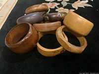 Photo showing eight vintage wooden bangles in various colors and shapes gathered in a group on a floral carpet. Batched display of different bangle styles and finishes visible.