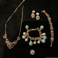 Photo showing vintage costume jewellery set on black background including gold-tone necklace with purple stones, pair of clip-on blue and amber stone oval earrings, a bracelet with floral and amber stones, and a charm bracelet with blue and pearl-like charms. American quarter added for size reference.