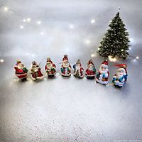 Front view of 8 distinct blown glass Santa Claus Christmas tree ornaments arranged in a line on a white surface with a lit Christmas tree in background.
