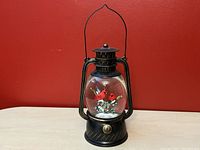 Front view of the black metal lantern snowglobe showing two red cardinal birds inside the clear glass globe, sitting on a light-colored surface against a red background.