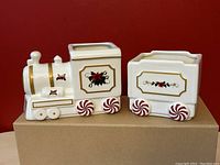 Front and side view of two-piece ceramic Christmas train candy dish set, showing detailed design on white ceramic with gold trim and red peppermint wheels.