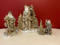 Group of three ceramic golden Santa figurines and three gold pine tree decorations on wooden surface against red background
