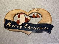 Wooden hand-painted Merry Christmas sign with Santa and bells on a heart-shaped plaque, shown from front view on a neutral background.