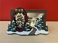 Front view of the ceramic Christmas decoration showing the oval frame with Christmas tree and snowy village backdrop.