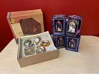 Lot showing five vintage glass decorative bulbs in an open cardboard box and four boxed Sunsweet Animal Collect-A-Bell ornaments standing upright behind the box against a red wall.