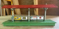 Green base with red roof railway station platform accessory with various vintage fuel advertising signage. Used as a train or railway accessory.