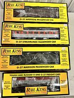 Four Rail King model train cars in original boxes showing branded packaging, model names, and visible train car details through clear plastic windows.