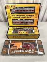 Stacked Rail King branded packages showing the O-27 Streamlined Passenger Combine/Diner 2-Car Set and two Rugged Rails series freight cars.