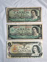 Three Canadian one dollar bills from 1954, 1967, and 1973 displayed on fabric showing front faces with Queen Elizabeth II and clear serial numbers.