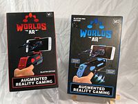 Front view of two boxed Worlds AR augmented reality game controllers, showing red and blue blasters with smartphone holders and branding.