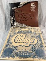 Two Chicago LP vinyl records with distinct covers; one brown with foil tear embossed logo, one cream with blue ornate design.