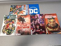 Top view of five DC Comics comic books arranged side by side on a surface. Titles include Steel, I Am Batman, and Superman: Worlds of War among others.