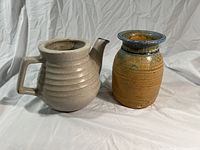 Photo showing a gray ribbed pottery jug and a brown glazed pottery tea pot side by side on a white fabric background.