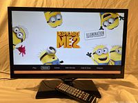 Front view of Samsung 23-inch LED TV showing screen powered on with animated Despicable Me 2 menu screen and remote control at base.