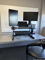 Front view of the standing desk converter on a white table, featuring two mounted monitors, laptop, keyboard on blue retractable tray, and mouse, with standing chair in foreground.