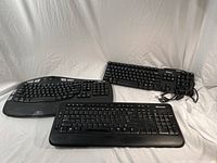 Three keyboards placed on white fabric background, showing Logitech wireless ergonomic keyboard, Dell wired keyboard with cable, and Microsoft wired keyboard with cable.