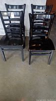 Four black wooden ladder-back chairs arranged in two rows on a concrete floor, visible scratches and wear on paint finish.