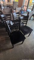 Four black wooden ladderback chairs with horizontal slats on backrest, showing scratches and wear on seats and frames, arranged on concrete floor in a warehouse setting.