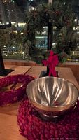 Photo of one green Christmas wreath decorated with a red bow, a deep red wreath, and a large stainless steel bowl on a wooden surface near a window at night.