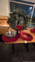 Green Christmas wreath with pinecones and a red bow, two red wreaths, and stainless steel bowl on a wooden table.