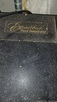 Close-up photo of the Excalibur logo on the dehydrator surface showing 'Excalibur Food Dehydrator' in gold lettering on a textured black background.