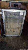 Front view of the metal industrial food warmer with closed glass door, showing temperature gauge on left side and three wire racks inside.