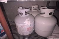 Photo showing three white propane tanks on metal shelving with one tank having a visible safety label
