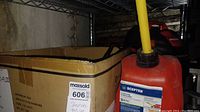 Photo showing red 2-gallon gas can with yellow spout and a cardboard box beside it on metal shelving.