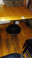 Full view of wooden table top with natural finish and round black metal base on wooden floor. Pizza boxes stacked nearby.