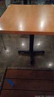 Full view of wooden square table top with metal black pedestal base on concrete floor, showing surface scratches