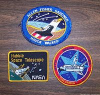 Top-down photo showing all three embroidered NASA mission patches on a wooden surface, clearly depicting the Shuttle and astronaut names, Hubble patch, and Columbia patch.