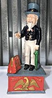 Front view of painted cast iron Uncle Sam coin bank standing on base with eagle emblem and briefcase.