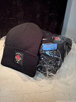 Lot includes 10 black knitted toques, one shown out of plastic packaging with a patch on the cuff featuring a red and blue design; the rest are still in the plastic bag.