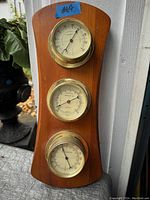 Set of three brass mounted weather instruments vertically arranged on a wood plaque shown in natural lighting.