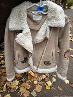 Front left side view of beige suede bomber jacket with white faux shearling collar and trim, front zipper partially open, decorative patches visible on front and sleeve.