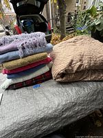 Photo showing stack of six assorted blankets next to folded double brown comforter on table outdoors.
