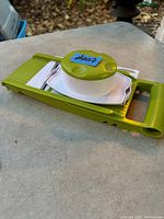 Green plastic mandolin slicer on a table, showing handle, straight blade, and white circular food holder with prongs.