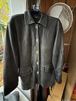 Front view of black leather coat hanging showing full length, collar, buttons, and pockets.