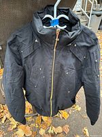 Front view of black Moose Knuckles men's winter jacket displayed on hanger outdoors showing zippered pockets, snap button details, and gold zipper.
