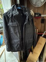 Black men's winter jacket hanging on a door, showing front with multiple large pockets and hood.