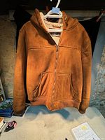 Front view of men's brown winter jacket with hood, showing zipper closed, front pockets, ribbed cuffs, and waistband on table.