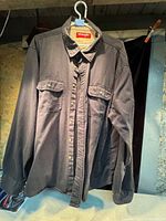 Photo shows a men's Wrangler long sleeve cotton shirt hanging on a white hanger, displaying button front and two chest pockets with buttons.
