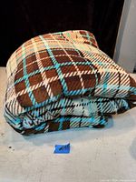 Folded large blanket with a brown, white, and light blue plaid pattern, shown on a flat surface.
