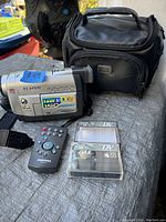 Photo of Samsung camcorder, remote, MiniDV tapes, and carrying case on a gray surface outdoors.