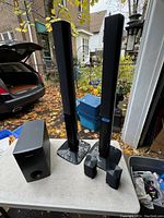 Photo of two tall Samsung floor-standing speakers, two smaller satellite speakers, and a subwoofer on a table outside.