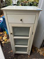 Front view of the small white cabinet showing drawer and glass door with shelves inside.