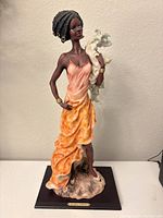 Front view of African woman sculpture wearing orange dress and holding white florals, standing on naturalistic base with Montefiori collection label.